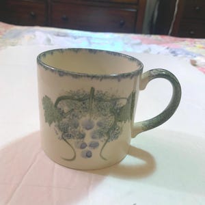 May include: Cream-colored ceramic mug with a green and blue grape design. The mug has a rounded handle and a blue-green trim around the rim. The grape design features green leaves and blue grapes.