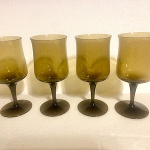 May include: Four vintage wine glasses with a unique, flared shape. The glasses are a warm amber color with a subtle sparkle, and they sit on dark brown stems and bases. A set of elegant glassware.