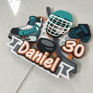 May include: A hockey-themed cake topper featuring a teal and white ice skate, a hockey helmet, a puck, and the number 30. The name "Daniel" is written in orange and white. The topper is on a clear stick.