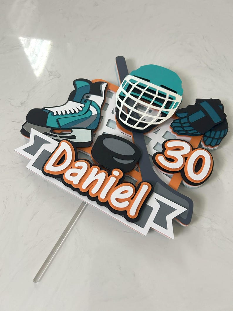 May include: A hockey-themed cake topper featuring a teal and white ice skate, a hockey helmet, a puck, and the number 30. The name "Daniel" is written in orange and white. The topper is on a clear stick.