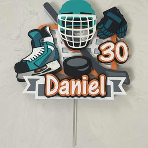 May include: A hockey-themed cake topper featuring a teal helmet, skate, and gloves, along with a hockey stick, puck, and the number 30. The name "Daniel" is displayed in orange and white text on a gray banner.