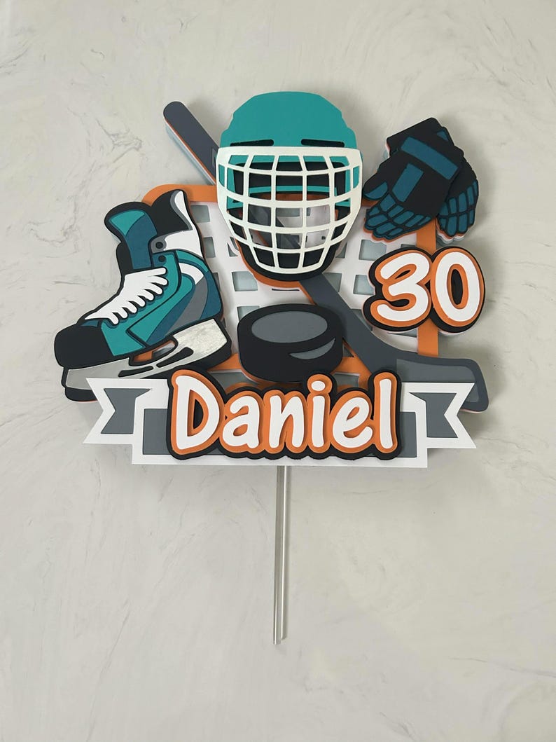 May include: A hockey-themed cake topper featuring a teal helmet, skate, and gloves, along with a hockey stick, puck, and the number 30. The name "Daniel" is displayed in orange and white text on a gray banner.