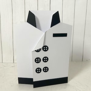 May include: A white paper box shaped like a suit jacket, with black accents. The box features six black buttons, a black collar, and a black pocket detail. The design mimics a formal jacket.