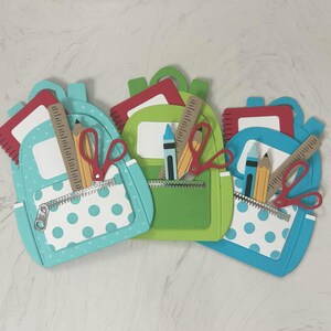May include: Three colorful paper cut-out backpacks in blue, green, and teal, each with a white polka-dot pocket. Each backpack contains a ruler, pencil, crayon, and scissors, suggesting a back-to-school theme.