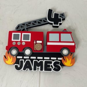 May include: A red and black fire truck cake topper with the name "JAMES" and the number "4". The topper features a ladder, flames, and a detailed fire truck design. Perfect for a birthday celebration.