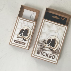 May include: Two open gift boxes with skull designs. One box has the text "A Wicked Thing" and the other says "Wicked". The boxes are brown with white and tan skull cutouts. One box contains a gift card.