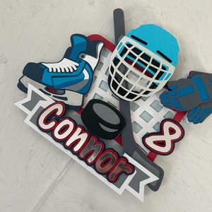 May include: A layered hockey-themed cake topper featuring a blue ice skate, hockey helmet, gloves, and a hockey stick. The name "Connor" is displayed in red and white lettering, along with the number "8".