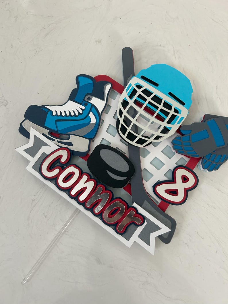 May include: A layered hockey-themed cake topper featuring a blue ice skate, hockey helmet, gloves, and a hockey stick. The name "Connor" is displayed in red and white lettering, along with the number "8".