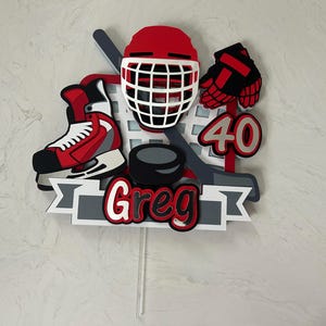 May include: A hockey-themed cake topper featuring a red helmet, skate, and gloves, along with a hockey stick and puck. The topper includes the name "Greg" and the number "40" in red and white. The design is set against a gray and white banner.