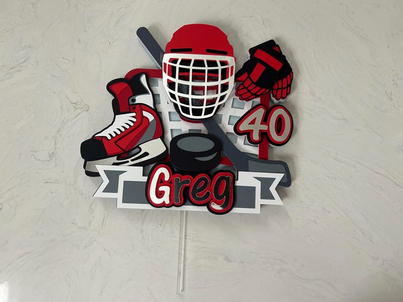May include: A hockey-themed cake topper featuring a red helmet, skate, and gloves, along with a hockey stick and puck. The topper includes the name "Greg" and the number "40" in red and white. The design is set against a gray and white banner.