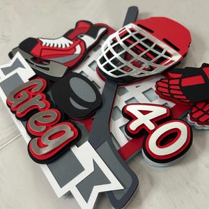 May include: A layered hockey-themed cake topper with a red, black, and white color scheme. The design includes a hockey skate, helmet, puck, stick, and the name "Greg" in silver and red, along with the number "40".