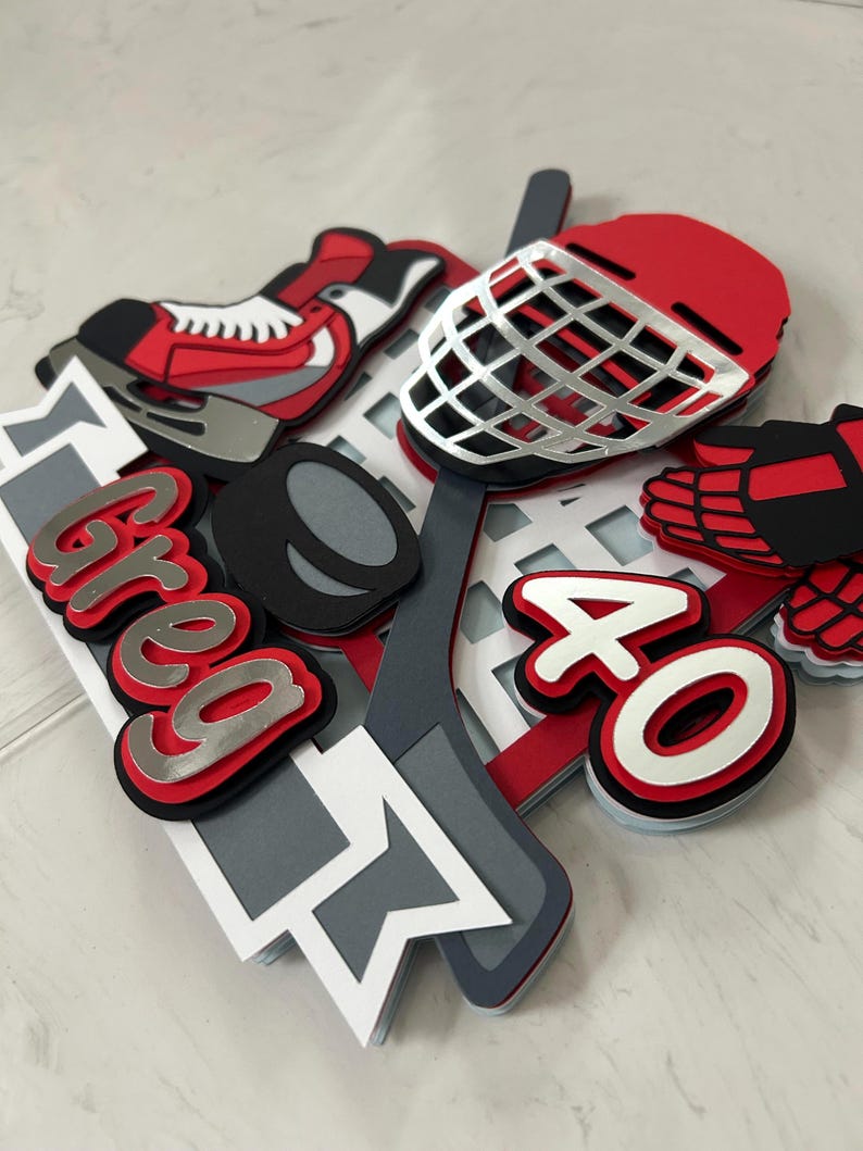 May include: A layered hockey-themed cake topper with a red, black, and white color scheme. The design includes a hockey skate, helmet, puck, stick, and the name "Greg" in silver and red, along with the number "40".