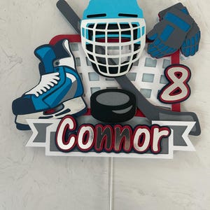 May include: A hockey-themed cake topper featuring a blue helmet, skates, gloves, and a hockey puck. The number 8 and the name "Connor" are also included. The design incorporates red, white, blue, and gray colors.