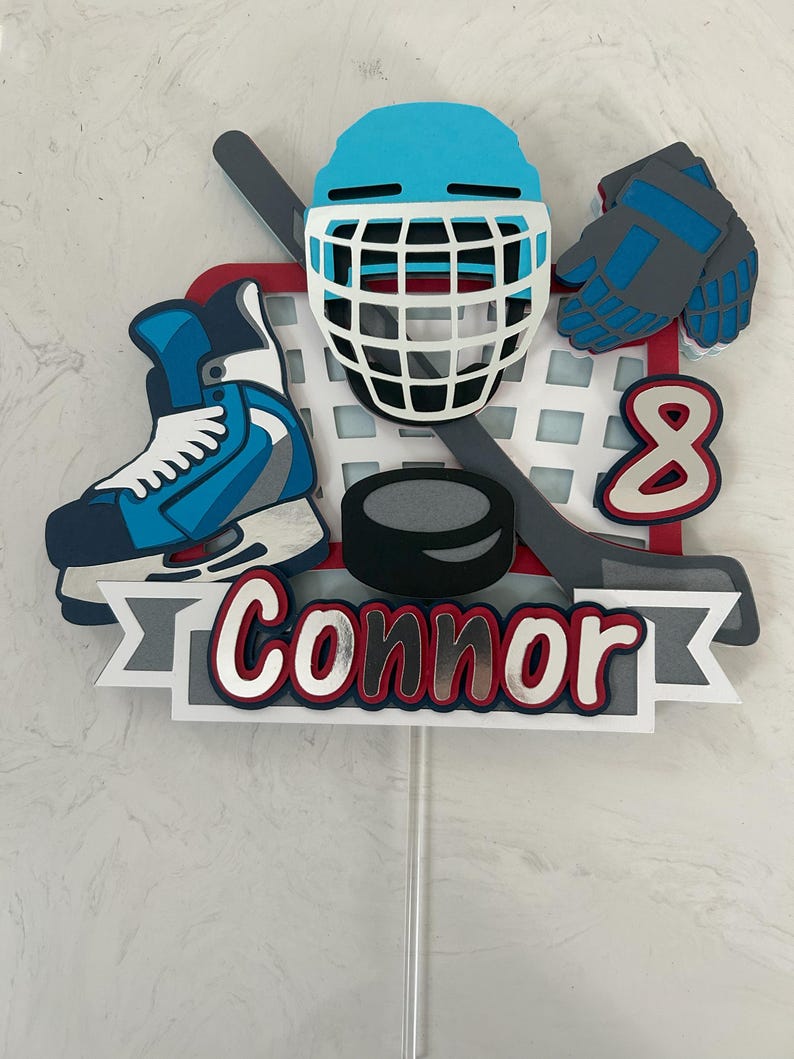 May include: A hockey-themed cake topper featuring a blue helmet, skates, gloves, and a hockey puck. The number 8 and the name "Connor" are also included. The design incorporates red, white, blue, and gray colors.