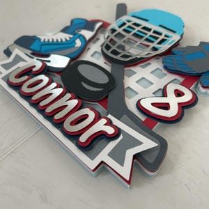May include: A layered paper hockey-themed decoration with a blue and white hockey skate, a blue helmet, a hockey puck, and the name "Connor" in red and silver lettering. The number "8" is also included.