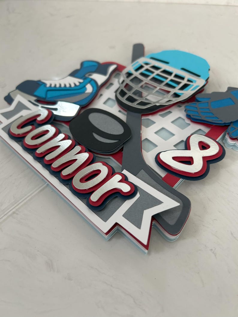 May include: A layered paper hockey-themed decoration with a blue and white hockey skate, a blue helmet, a hockey puck, and the name "Connor" in red and silver lettering. The number "8" is also included.