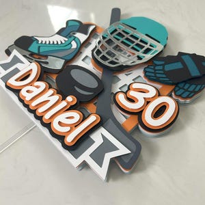 May include: A hockey-themed cake topper featuring a teal and silver hockey skate, helmet, and puck. The topper includes the name "Daniel" in orange and white, along with the number "30". The design is layered with orange, gray, and white accents.