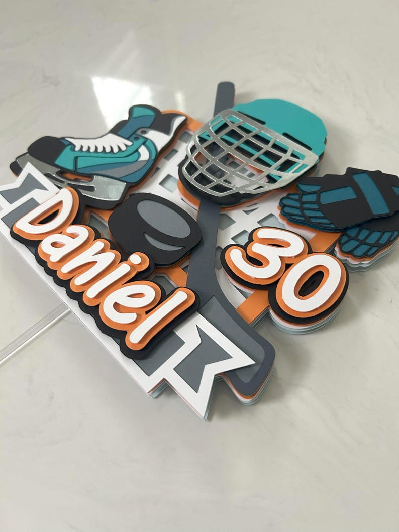May include: A hockey-themed cake topper featuring a teal and silver hockey skate, helmet, and puck. The topper includes the name "Daniel" in orange and white, along with the number "30". The design is layered with orange, gray, and white accents.