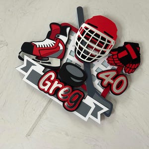 May include: A hockey-themed cake topper featuring a red and black hockey skate, helmet, and glove. The design includes a hockey puck, stick, and the name "Greg" and the number "40" in red and white.
