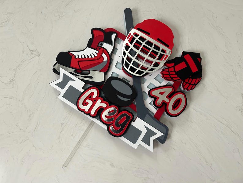May include: A hockey-themed cake topper featuring a red and black hockey skate, helmet, and glove. The design includes a hockey puck, stick, and the name "Greg" and the number "40" in red and white.