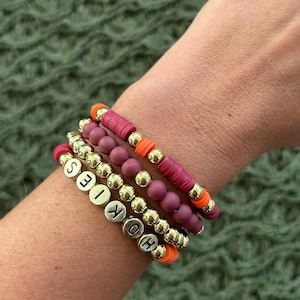 May include: A set of four bracelets featuring gold beads, burgundy beads, and orange beads. The bracelets spell out the word "HOKIES" in gold letters.