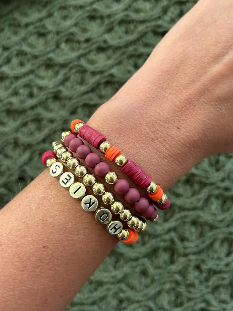 May include: A set of four bracelets featuring gold beads, burgundy beads, and orange beads. The bracelets spell out the word "HOKIES" in gold letters.