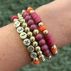 May include: Four stacked bracelets with gold beads and red and orange accents. The top bracelet has the word "HOKIES" spelled out in black letters.