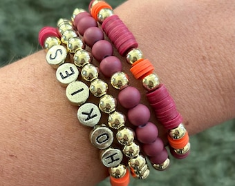 May include: Four stacked bracelets with gold beads and red and orange accents. The top bracelet has the word "HOKIES" spelled out in black letters.