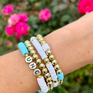 May include: A stack of bracelets with gold beads and white and light blue spacer beads. The top bracelet has the letters "UNC" spelled out in silver beads.