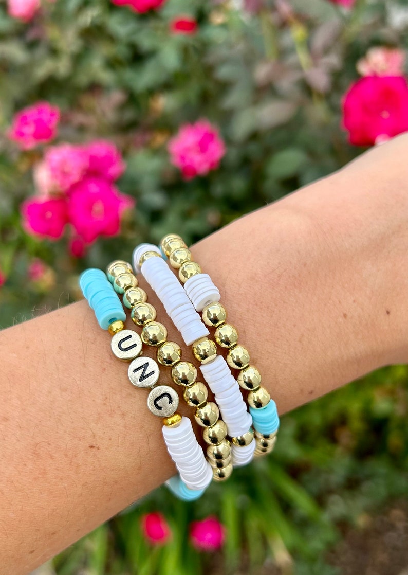 May include: A stack of bracelets with gold beads and white and light blue spacer beads. The top bracelet has the letters "UNC" spelled out in silver beads.