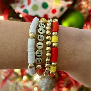 May include: A set of four bracelets, one with white beads, one with gold beads, one with red and yellow beads, and one with gold beads and the word "CHIEFS" spelled out in black letters.