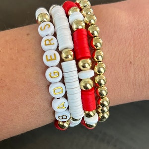 May include: A set of four bracelets with white beads that spell out "BADGERS" in gold letters. The bracelets also feature red and gold beads.
