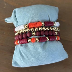May include: A set of five bracelets featuring maroon, white, and orange beads. The bracelets are stacked on top of each other and are displayed on a light blue fabric background.