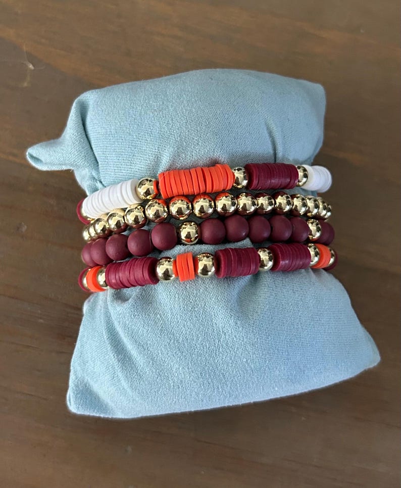 May include: A set of five bracelets featuring maroon, white, and orange beads. The bracelets are stacked on top of each other and are displayed on a light blue fabric background.