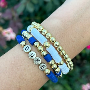 May include: A set of four bracelets with gold beads and blue and white spacers. The bracelets spell out the word "DUKE" in black letters.