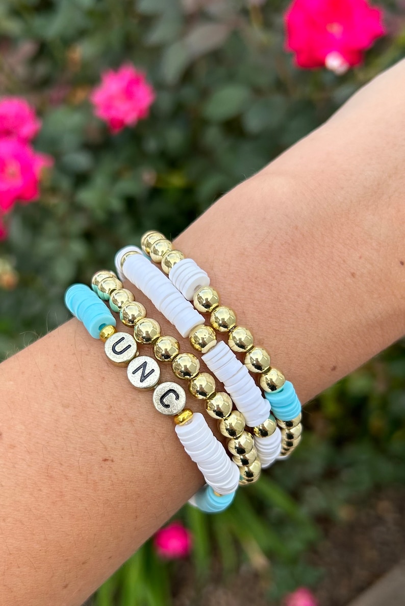 May include: A set of four bracelets with gold beads and white and turquoise spacers. The bracelets spell out the word "UNC" in silver letters.