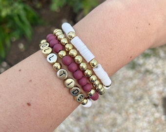 May include: A set of four bracelets featuring gold beads, white beads, and maroon beads. The bracelets spell out "ROLL TIDE" in gold letters.