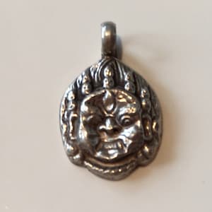 Könnte beinhalten: Ein silberfarbener Anhänger mit einem detaillierten, stilisierten Gesicht mit einem grimmigen Ausdruck. Der Anhänger hat oben eine Schlaufe für eine Kette und ist wahrscheinlich aus Metall. Das Gesicht weist komplizierte Details auf, darunter eine prominente Stirn und ein breiter Mund.