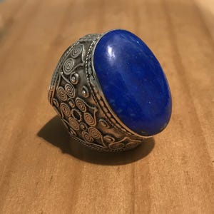 Può includere: Un anello d'argento con una pietra ovale di lapislazzuli. L'anello presenta un design dettagliato di motivi a spirale.