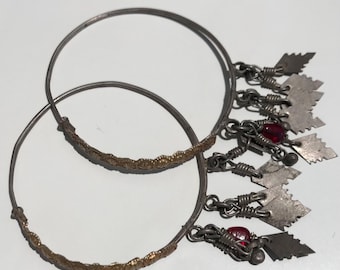 Könnte beinhalten: Zwei silberfarbene Creolen mit goldfarbenen Details. Jeder Ohrring hat baumelnde Anhänger, darunter kleine silberne Blätter und rote Perlen. Die Ohrringe werden auf einer weißen Oberfläche präsentiert.