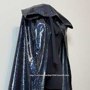 May include: A black, iridescent cape with a large hood. The cape has a shiny, textured surface that reflects multiple colors. The cape is draped over a black garment. The Etsy shop URL is visible at the bottom.
