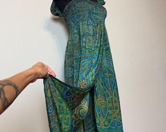 May include: A long, flowing dress with a vibrant paisley pattern in shades of teal, green, and gold. The dress features a ruffled neckline and sleeves, and is displayed on a black mannequin. The fabric is held up by a hand.