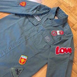 May include: An olive green button-down shirt adorned with various patches. Patches include the word "SETTERS", a red heart, a red and white "LOVE" patch, and other colourful designs. The shirt features two chest pockets and long sleeves.