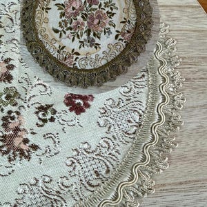 Può includere: Un centrino rotondo e una sezione di una tovaglia abbinata, entrambi con motivi floreali. Il centrino presenta un bouquet centrale di fiori rosa e bianchi con un bordo in pizzo marrone. La tovaglia ha uno sfondo color crema con motivi floreali e a scorrimento.