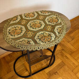 Può includere: Runner da tavolo ovale con motivo floreale nei toni dell'oro, del marrone e del crema. Il runner presenta un design esagonale con bouquet di fiori e un bordo in pizzo dorato decorativo. Il runner è posto su un tavolino rotondo.