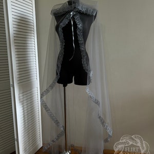 May include: A sheer silver cape with a lace trim and a ribbon tie. The cape is draped over a mannequin and is displayed against a white wall.