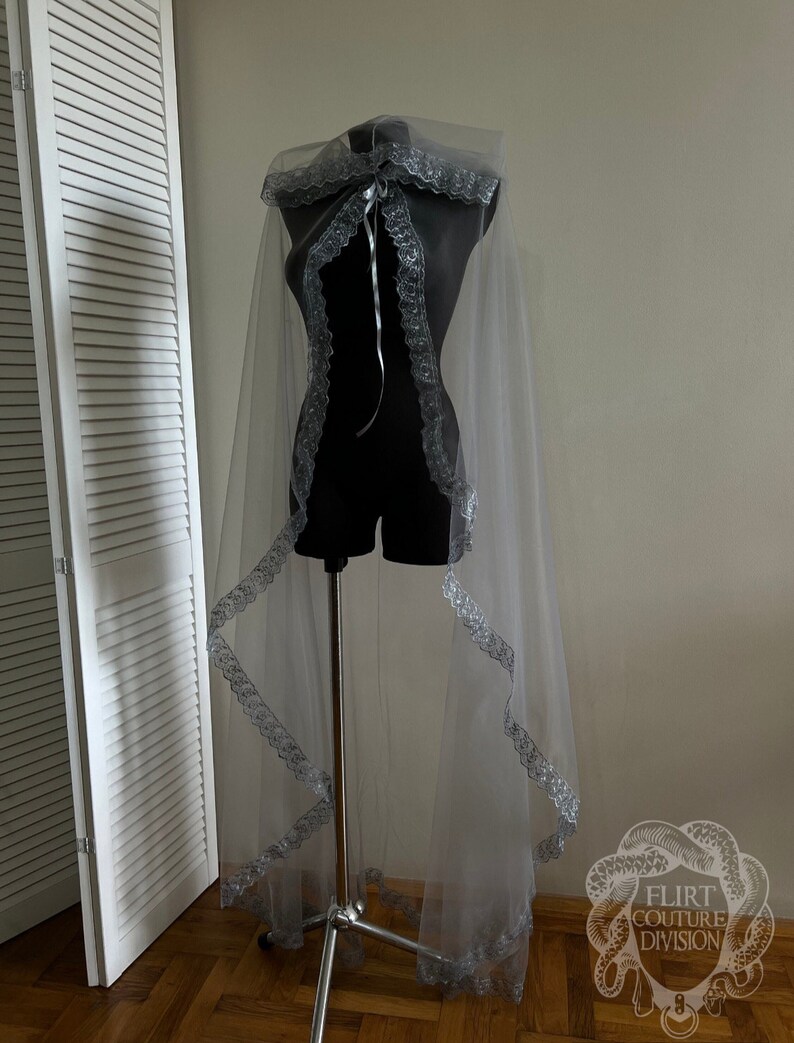 May include: A sheer silver cape with a lace trim and a ribbon tie. The cape is draped over a mannequin and is displayed against a white wall.