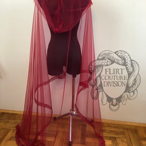 May include: A long, flowing burgundy tulle cape with a lace trim and a hood. The cape is draped over a dress form and the fabric is sheer and delicate.