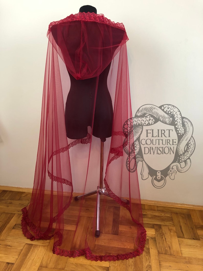 May include: A long, flowing burgundy tulle cape with a lace trim and a hood. The cape is draped over a dress form and the fabric is sheer and delicate.