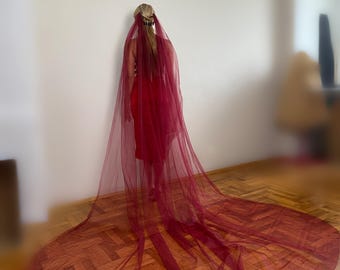 May include: A long, flowing burgundy veil, likely for a wedding or special occasion, trails across a wooden floor. The veil is sheer and drapes dramatically, creating a circular shape. The person is wearing a red dress.